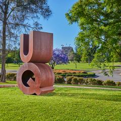 A bronze statue of the letters U and Q on a lawn in front of trees. The U is stacked on top of the Q.