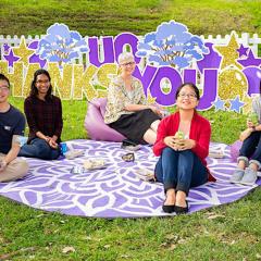 Five people sitting on a round picnic blanket in front of a low white fence and a big sign reading 'UQ Thanks You'