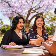 Three young adults sitting at a picnic table in front of a jacaranda tree